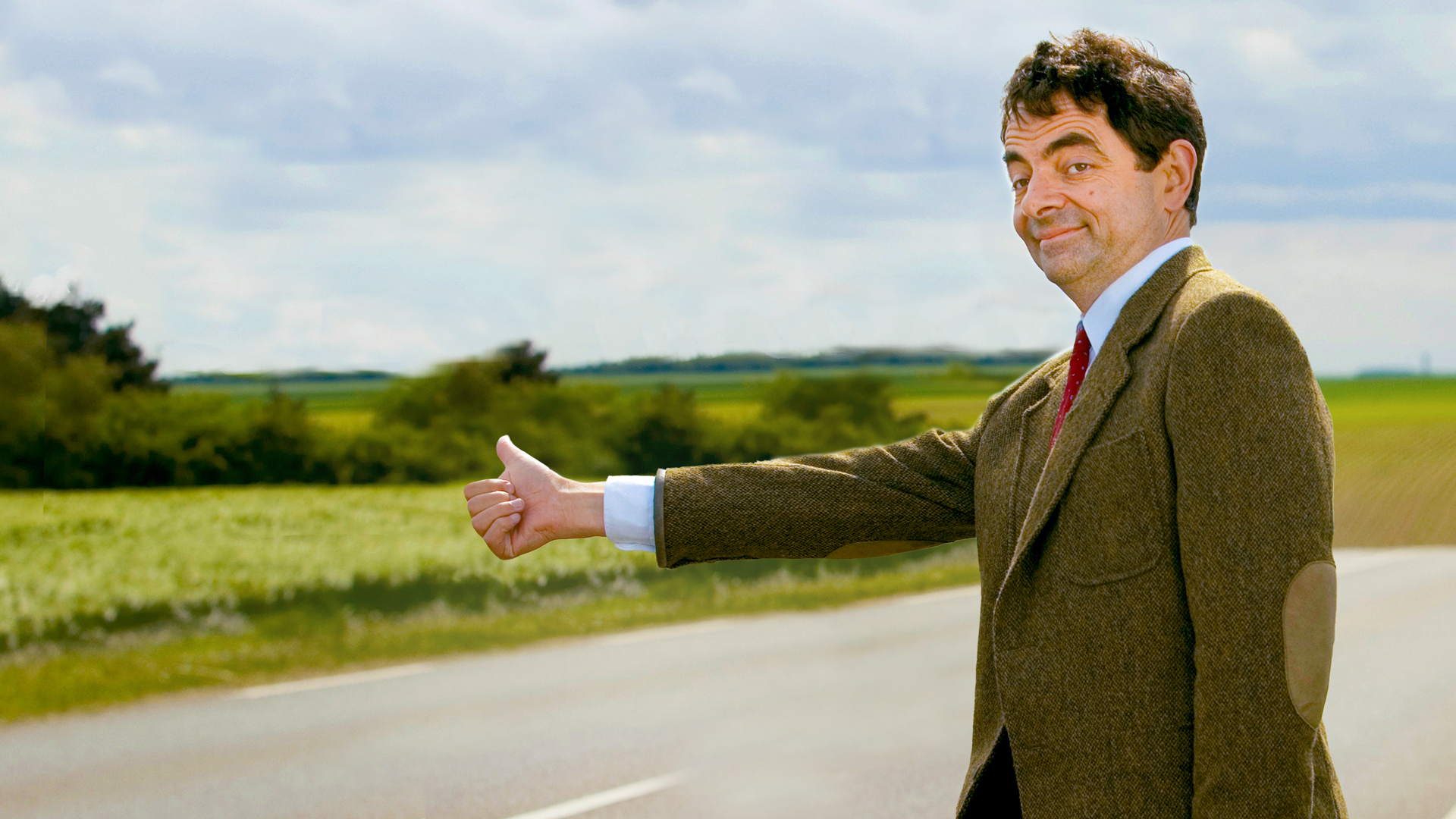 Mr. Bean steht mit ausgestrecktem Daumen am Straßenrand, im Anzug und mit schelmischem Blick. (Bild: rbb/ Studiocanal GmbH)