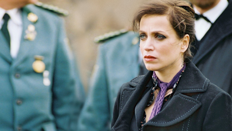Paula Merten, gespielt von Anna Thalbach, blingt mit trauriger Miene ins Leere. Im Hintergrund sind Menschen in Uniformen abgebildet.