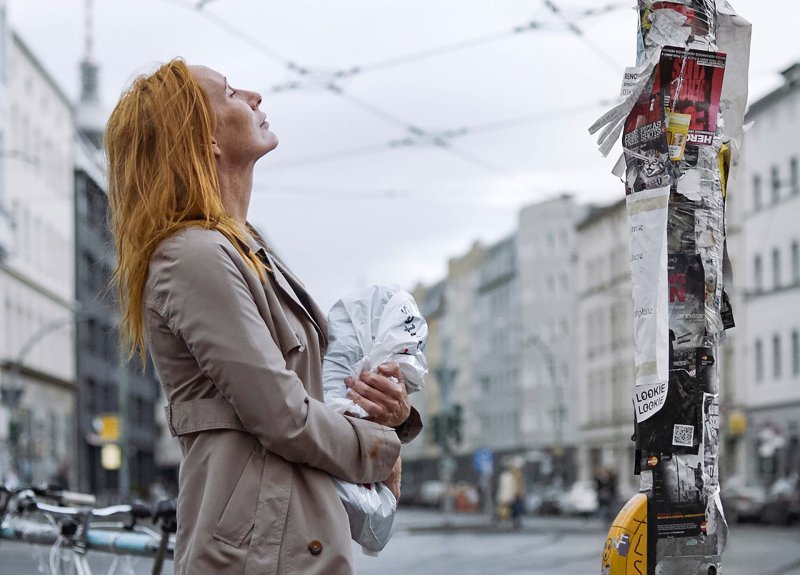Rothaarige Frau steht an einer Berliner Straßenecke, blickt nachdenklich in den Himmel und hält eine Plastiktüte in der Hand, neben ihr ein mit Zetteln und Plakaten beklebter Laternenpfahl.