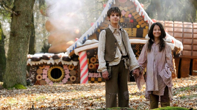 Ein ärmlich gekleidetes Geschwisterpaar hält sich an den Händen. Im Hintergrund ist ein großes Lebkuchenhaus zu sehen.