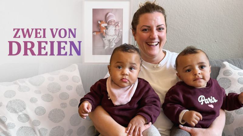 Teresa sitzt mit ihren beiden Zwillingsmädchen Zola und Zoey auf einer Coach, im Hintergrund ist ein gerahmtes Bild ihrer verstorbenen Tochter Zhara an der Wand zu sehen.