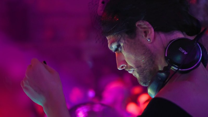 DJ im Profil mit Kopfhörern um den Hals, konzentriert am Mischpult; pink-violettes Clublicht, unscharfe Menge im Hintergrund.