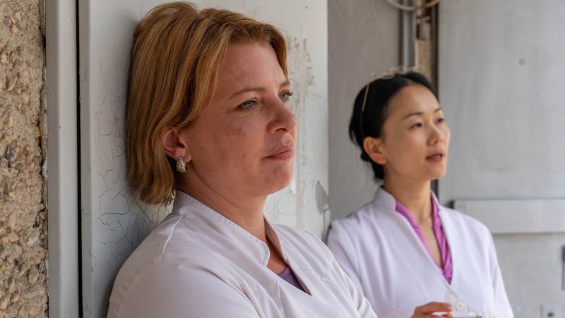 Kathi (Jördis Triebel) in weißem Kittel lehnt nachdenklich an einer Wand; im Hintergrund steht ihre Chefin Jenny (Yvonne Yung Hee Bormann).