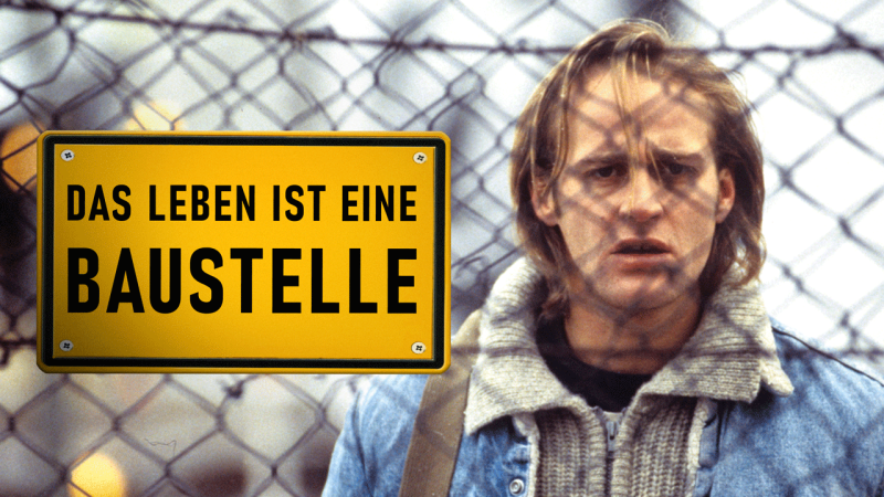 Schauspieler Jürgen schaut mit verdutztem Gesichtsausdruck durch einen Bauzahn an dem ein Schild mit der Aufschrift "Das Leben ist eine Baustelle" hängt. (Bild: NDR/BetaFilm) 