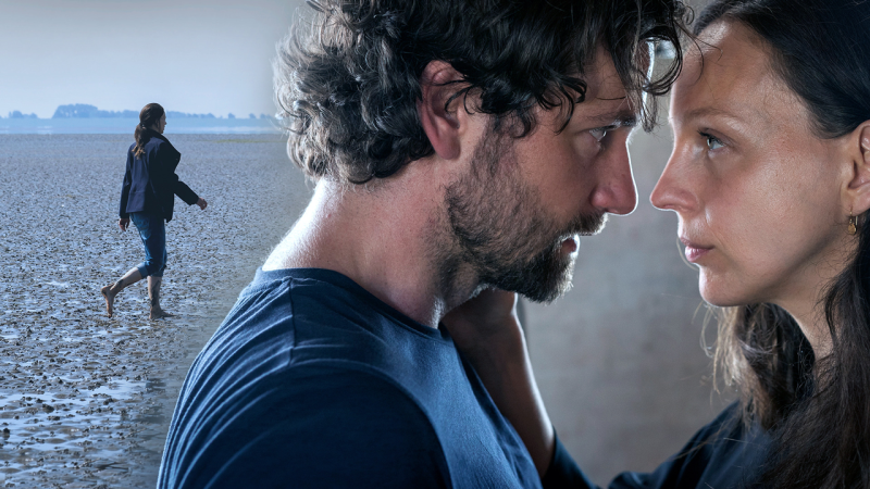 Florian Stetter und Petra Schmidt-Schaller sind einander zugewandt und schauen sich in die Augen, im Hintergrund spaziert eine Frau am Strand entlang. (Bild: ARD Degeto/Boris Laewen)ARD Degeto / Boris Laewen