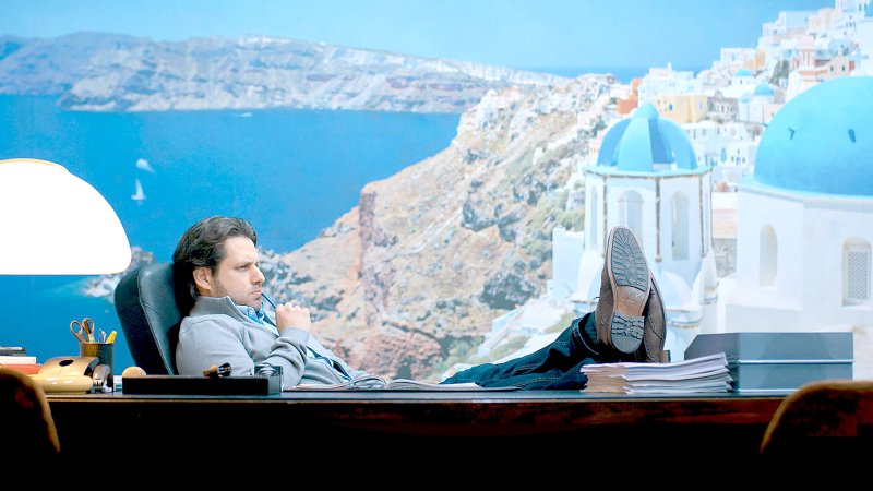 Dimitrios Schulze am Schreibtisch vor einer Fototapete mit griechischer Landschaft | Bild: SWR/kurhaus production / Johannes Krieg