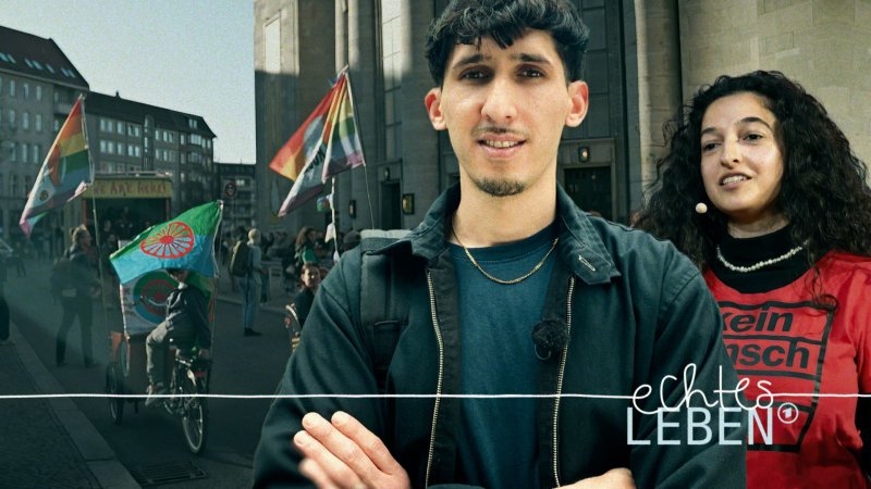 Ein junger Mann und eine junge Frau stehen vor einem Gebäude; im Hintergrund sind Menschen mit Roma- und Regenbogenflaggen zu sehen.