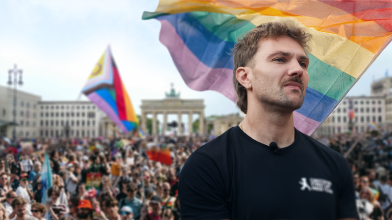 Ein junger Mann schaut mit angespanntem Gesichtsausdruck in die Ferne. Hinter ihm ist eine große Regenbogenflagge und eine Menschenmenge zu sehen.